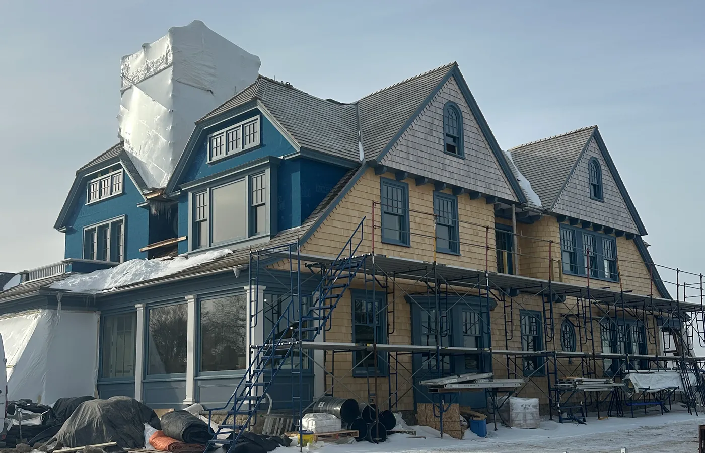 Historic shingle-style home with frame scaffolding and shrink-wrap containment on the chimney in winter